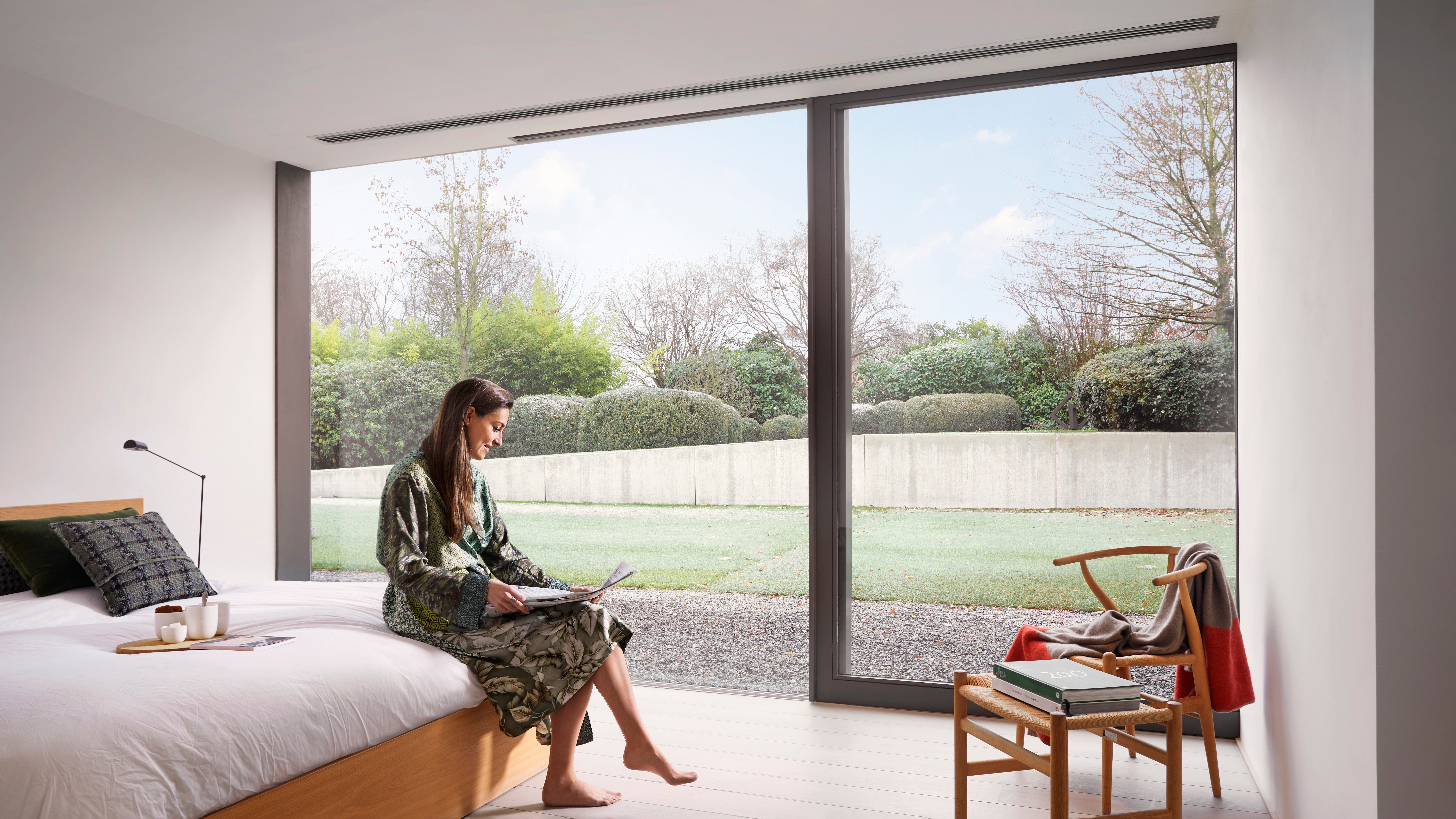 Een vrouw zit op de rand van haar bed de krant te lezen met haar ochtenddrankje vóór haar, en het plafondinbouwmodel zorgt voor een comfortabele temperatuur in de slaapkamer