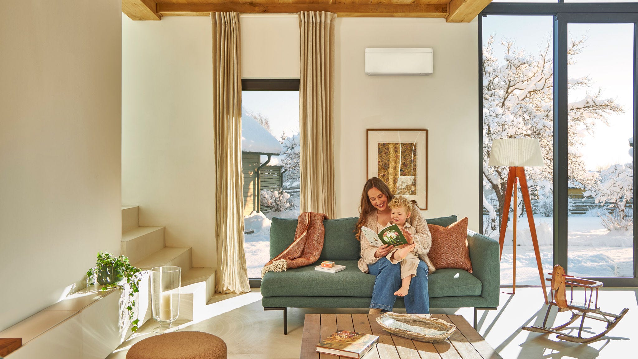 Moeder zit op de bank onder een lucht/lucht warmtepomp aan de wand en leest voor uit een boek voor haar kind dat op haar schoot zit. Ze genieten samen van de warmte terwijl het buiten sneeuwt.